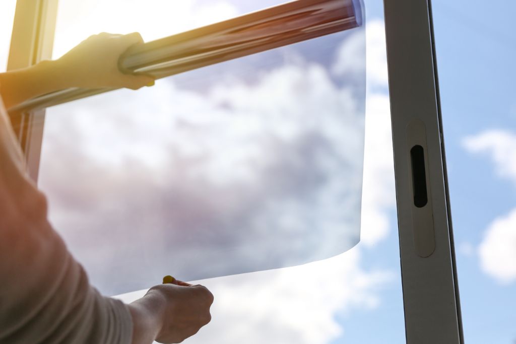 A close-up of someone applying window film to a glass window. The hands are holding the film while the sky with clouds is visible in the background.