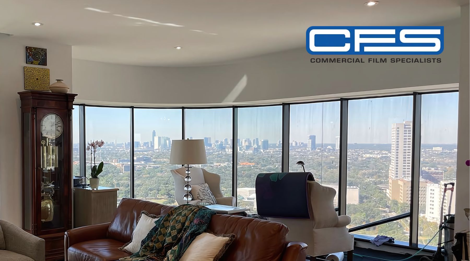 An interior shot of a high-rise apartment with floor-to-ceiling curved windows, offering a panoramic view of a city skyline. The room is stylishly furnished with a leather couch, a grandfather clock, and other modern decor elements. The "CFS" logo for Commercial Film Specialists is visible in the top right corner.