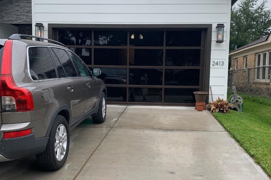 The image depicts the driveway of a modern home with a sleek, glass-paneled garage door. A gray SUV is parked to the left on the driveway. The house number "2413" is visible on the white exterior wall next to the garage door. On the right side of the garage, a small potted plant and a garden hose reel are placed near a patch of grass.