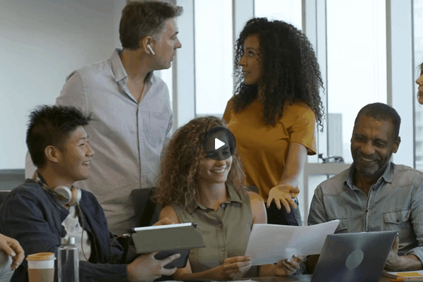 window tint Enhances Comfort and Productivity - This image shows a group of diverse professionals gathered in an office setting, smiling and engaging in conversation. They appear to be collaborating on a project, with one person holding papers and others using a laptop and tablet. The natural light from large windows suggests a bright workspace, potentially highlighting the role of window tinting in enhancing comfort and reducing glare in office environments.