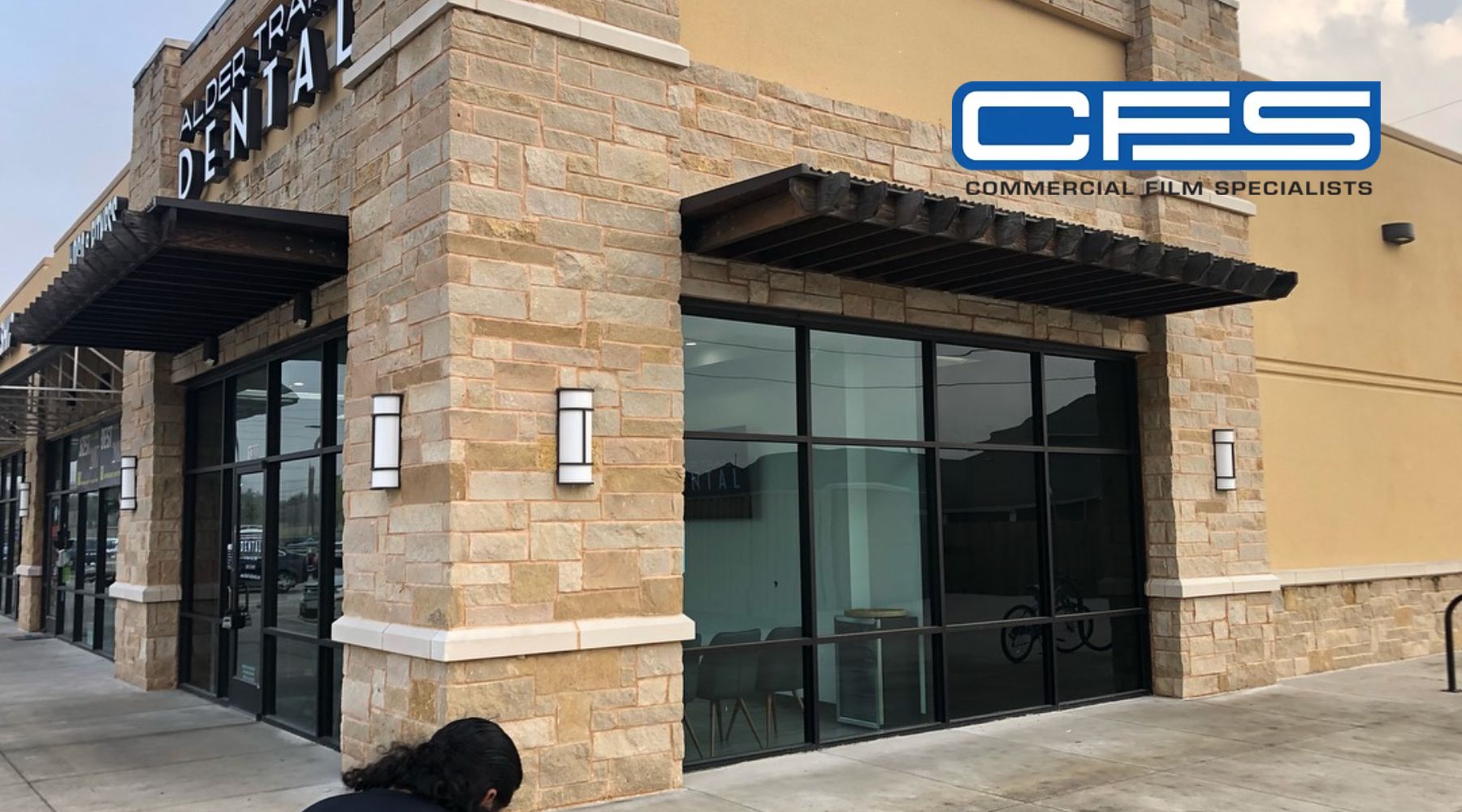 Benefits of Commercial Window Tinting - image shows the exterior of a commercial building with a sign reading "Alder Trails Dental." The building features stone siding and large glass windows, while the logo for "CFS Commercial Film Specialists" is displayed in the upper right corner. A person is crouching near the bottom left of the image, outside the building.