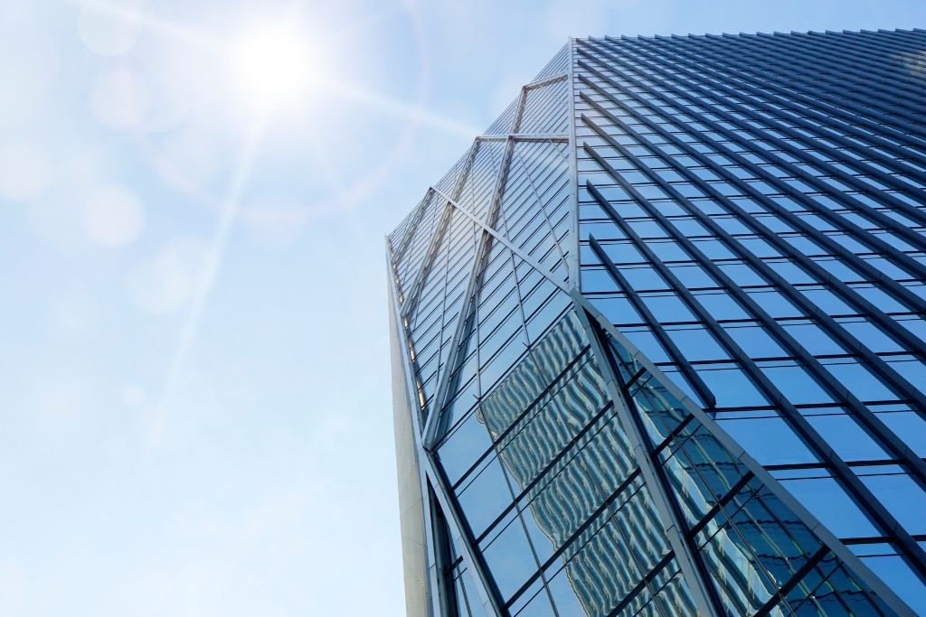 A tall, modern glass skyscraper reflecting the bright sunlight in the sky, with the sun shining directly onto the building's sleek surface.