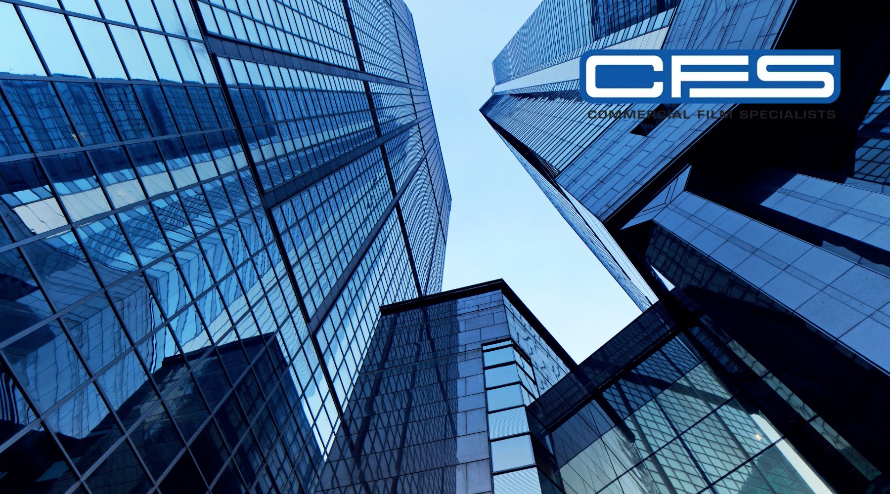 Looking up at modern glass skyscrapers reflecting the blue sky, with the 'CFS Commercial Film Specialists' logo in the upper-right corner. The image conveys a professional, sleek atmosphere, emphasizing architectural glass and window tinting services.