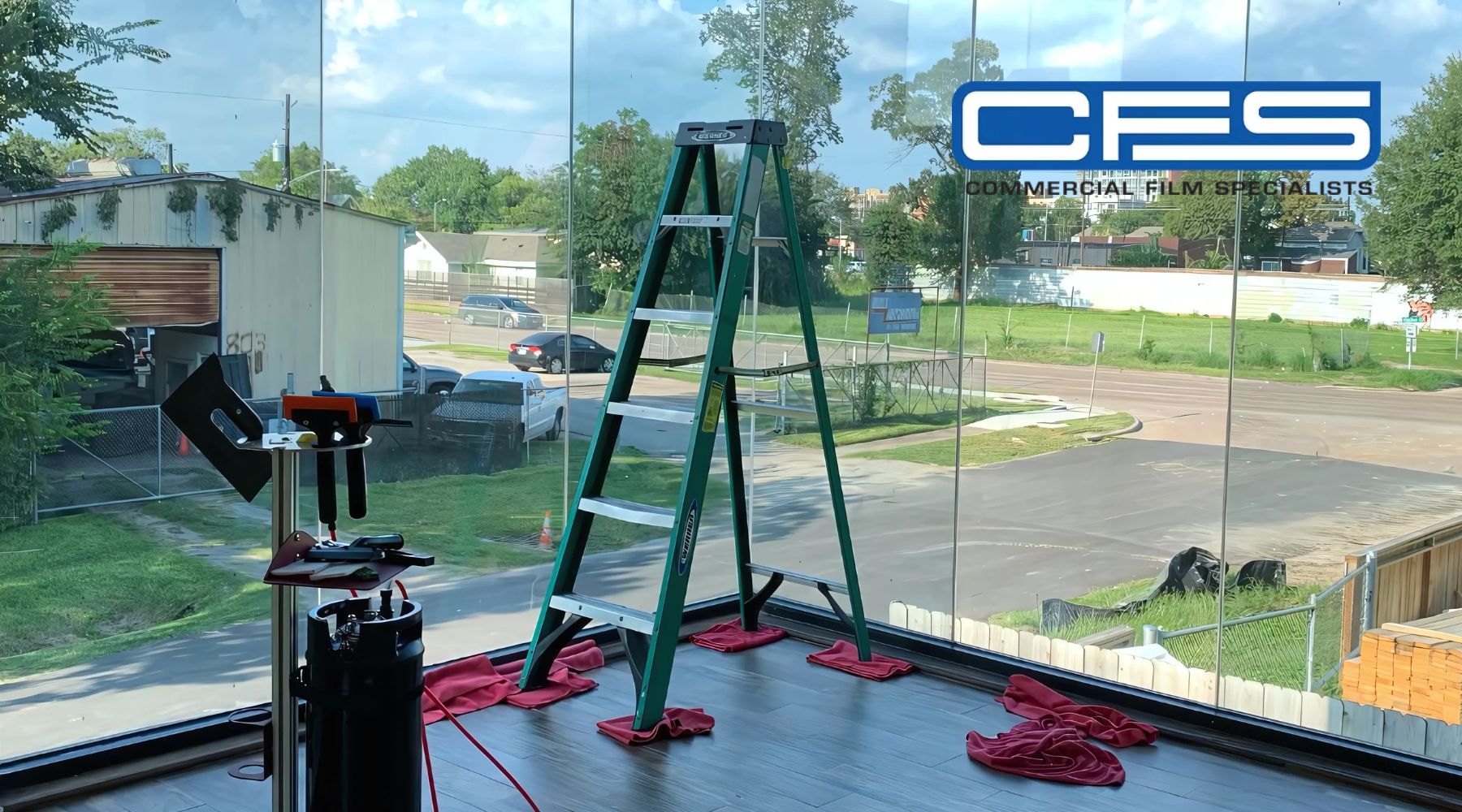 A green ladder stands near a large glass window, with red cloths positioned at its base, indicating a window installation or tinting project. Tools, including a squeegee and utility knife, are arranged on a nearby stand. The "CFS Commercial Film Specialists" logo is visible on the glass, hinting at professional involvement in the project.