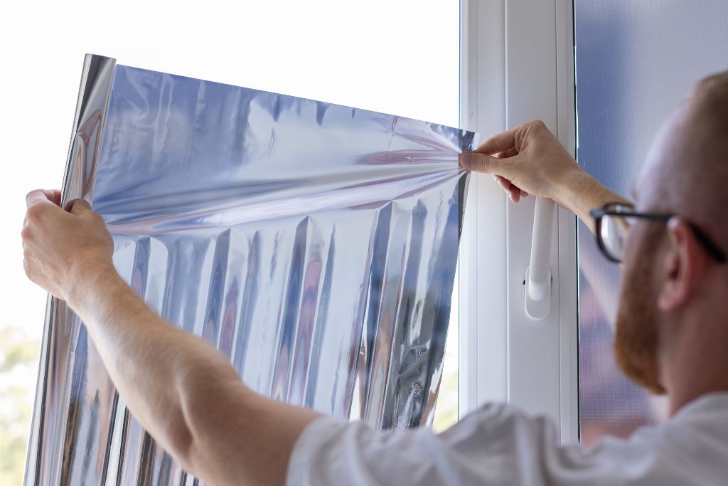 A person carefully applies a sheet of reflective window tint film onto a window, holding the film taut as they position it. The reflective film creates a mirrored effect, highlighting the precision needed in the DIY window tinting process.