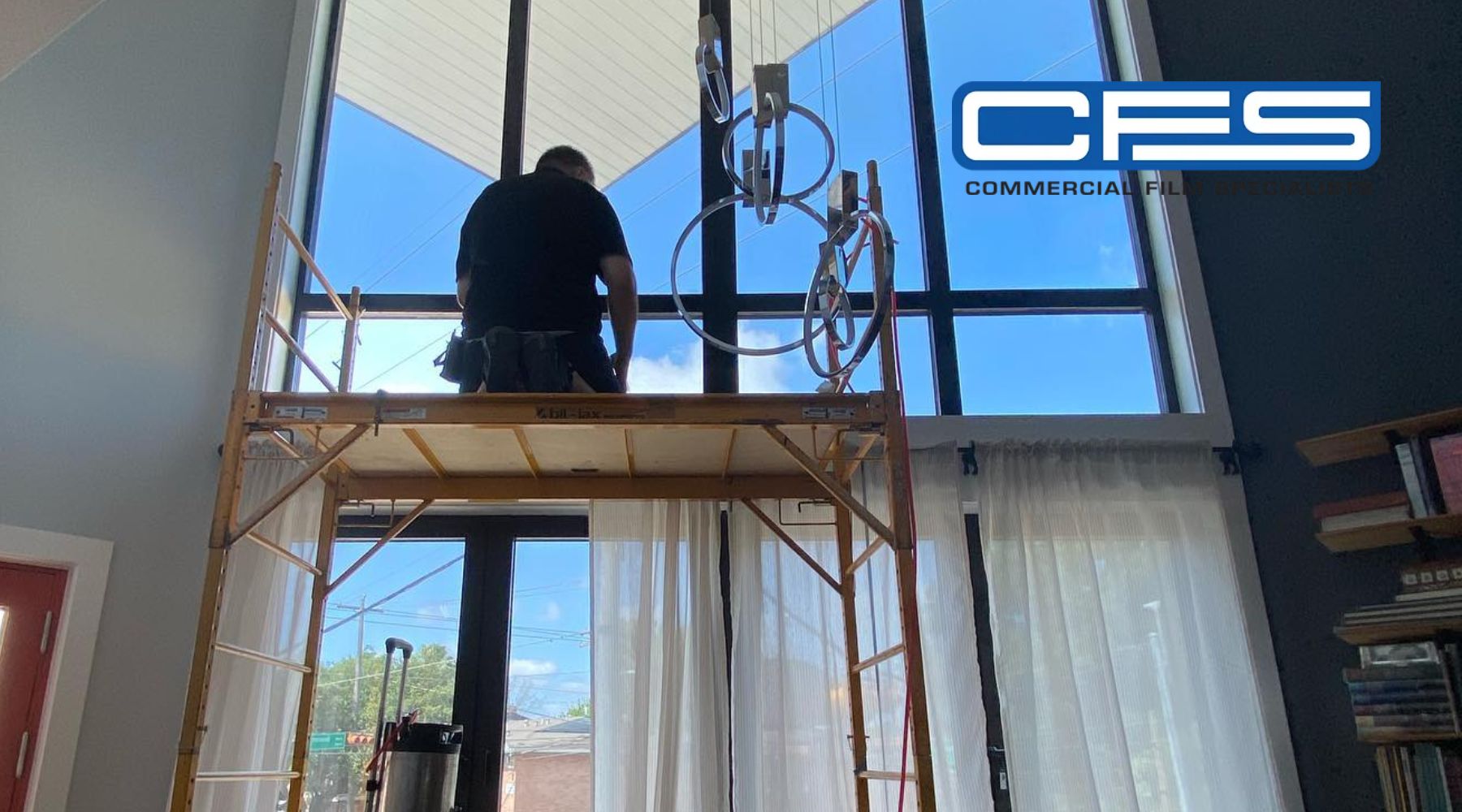 A technician from Commercial Film Solutions (CFS) is working on scaffolding inside a room with high, large windows, preparing to apply window tinting. The CFS logo appears in the top right corner, and the bright outdoor light contrasts with the interior's softer lighting.