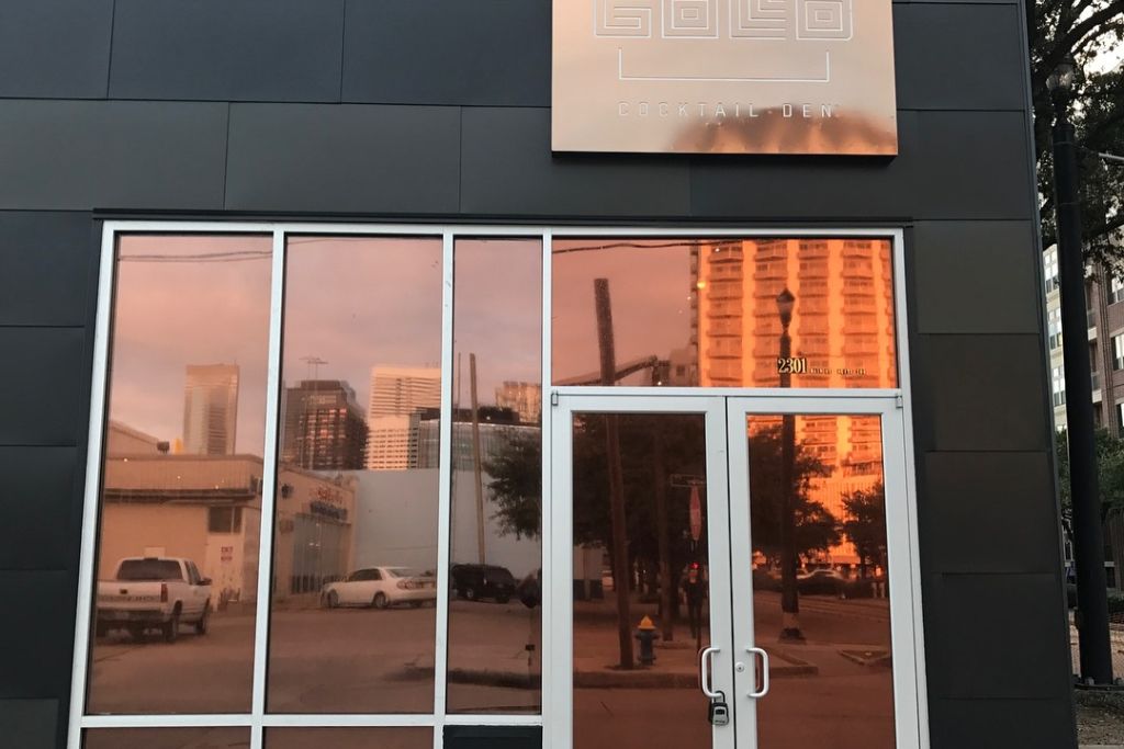 A storefront with reflective tinted windows showing a vibrant orange glow, mirroring the surrounding cityscape and skyline. The building features a modern black exterior with a sign reading “GOLD Cocktail Den” above the entrance.