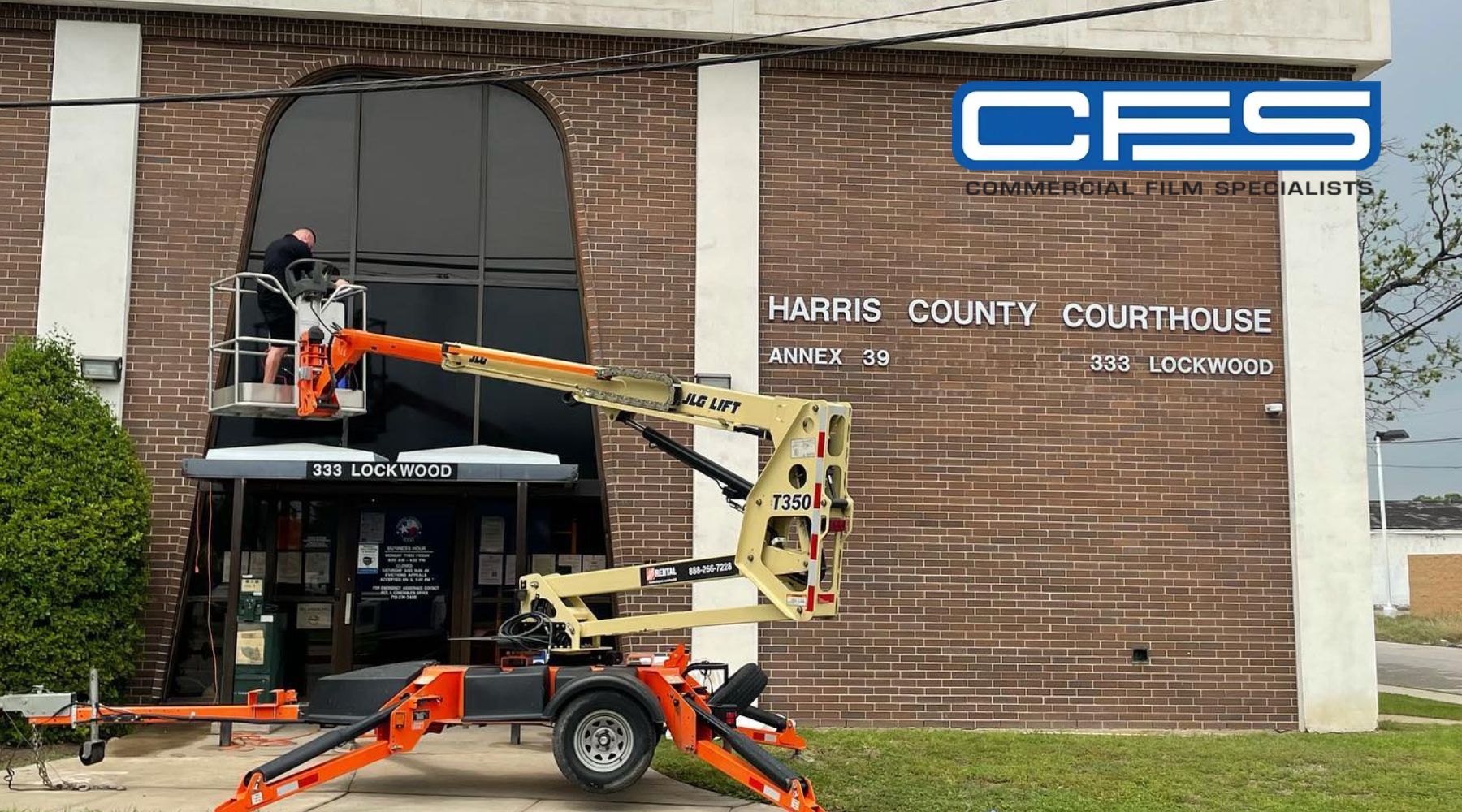 A worker on a mobile lift installs or maintains commercial window tinting on the large front window of the Harris County Courthouse Annex 39, located at 333 Lockwood. The building is made of red brick, with visible signage and the logo for CFS (Commercial Film Specialists) in the upper-right corner.