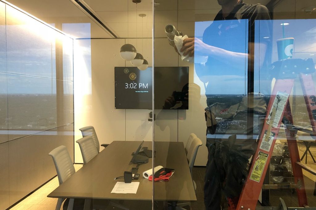 A modern office meeting room with glass walls, featuring a large table, chairs, and a mounted screen displaying the time “3:02 PM.” A technician is visible through the glass, working on window tinting installation while standing on a ladder, with a clear outdoor cityscape in the reflection.