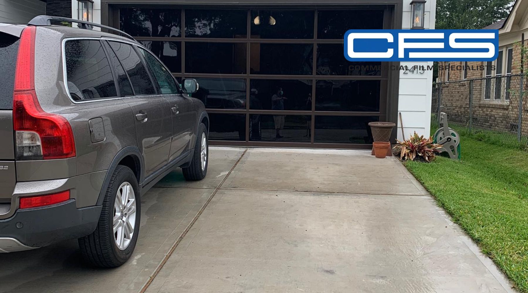 A gray SUV parked in front of a modern glass garage door, with the logo "CFS Commercial Film Specialists" prominently displayed in the upper right corner. The driveway and lawn appear wet from recent rain, and potted plants and a garden hose reel are visible near the side of the garage.