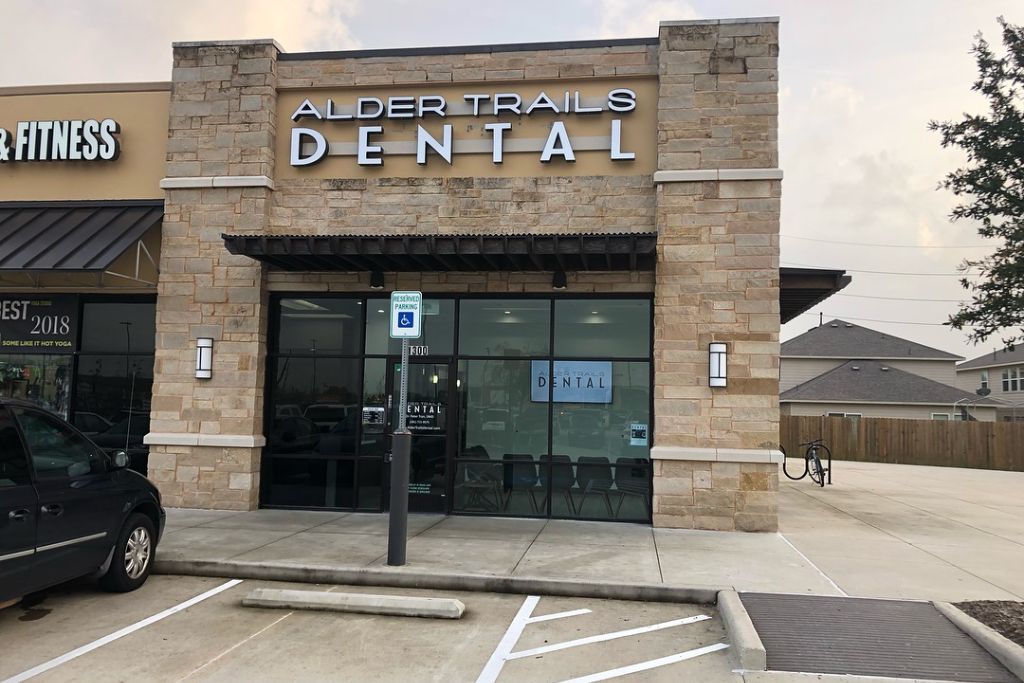 The storefront of Alder Trails Dental, a modern brick and stone building with large glass windows and a clear sign reading “Alder Trails Dental.” The entrance includes a handicapped parking space directly in front, and neighboring storefronts can be seen partially.