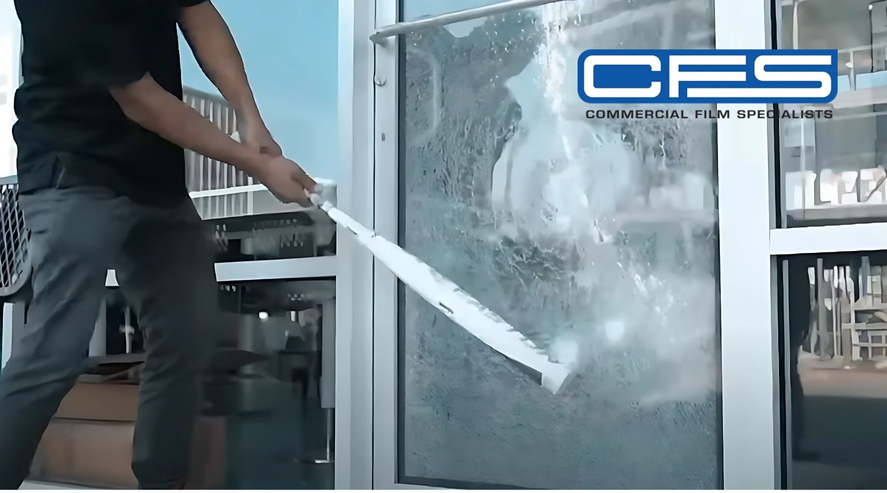 A person striking a glass window covered with protective film, showcasing the durability of commercial window films by CFS (Commercial Film Specialists). The partially shattered window demonstrates the film’s ability to hold the glass in place during impact.