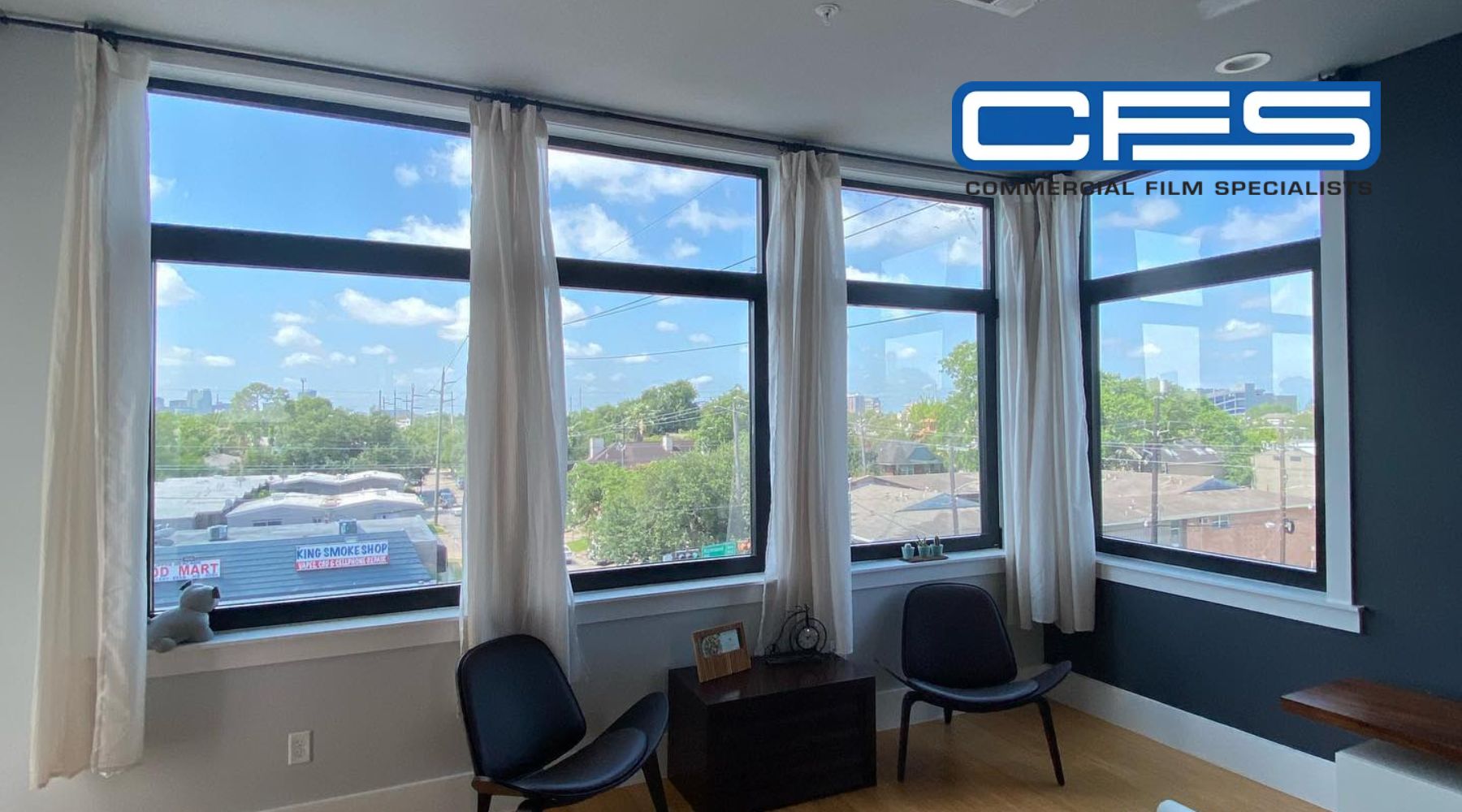 A modern interior space with large windows offering a clear view of the outdoors, featuring residential buildings and a commercial area. The windows are framed with white curtains, and the room is furnished with two black chairs and a small wooden table with decorative items. A "CFS Commercial Film Specialists" logo is displayed in the top right corner.