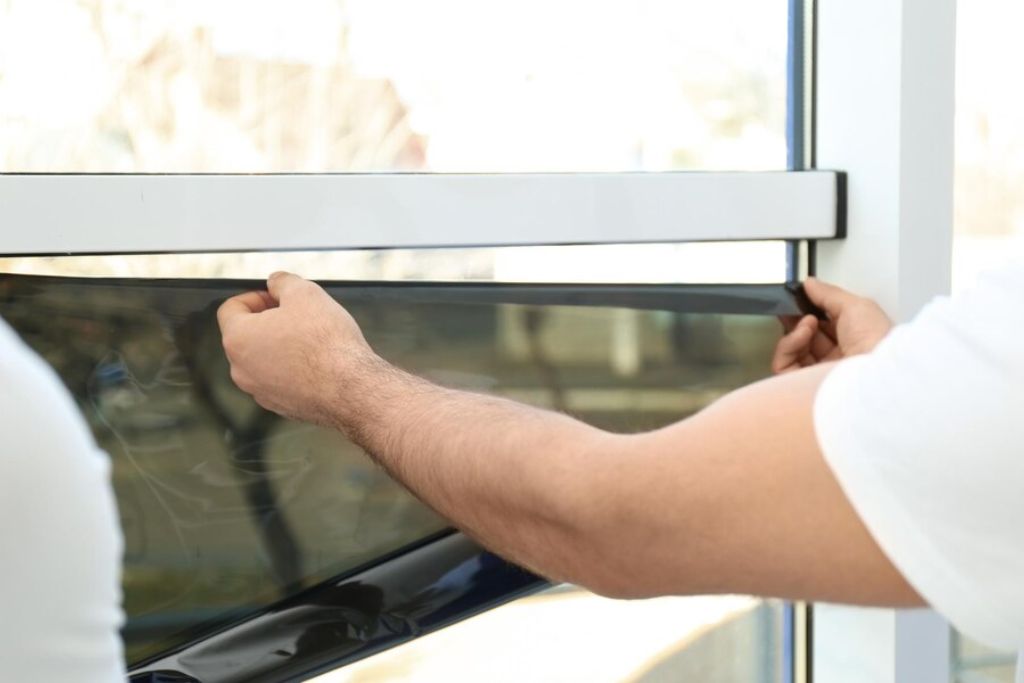 A professional installer carefully applies dark-tinted window film to a large glass window. The installer, wearing a white shirt, uses both hands to align the film precisely along the window frame to ensure proper placement. The window reflects outdoor scenery, and the film is partially smoothed out to eliminate air bubbles.