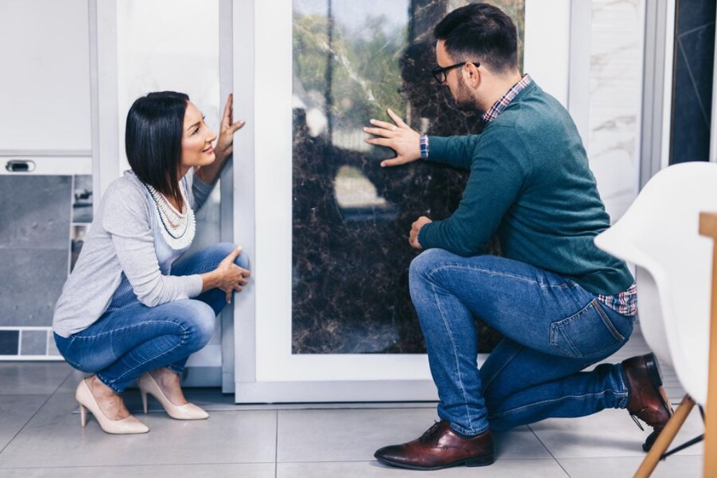 A man and a woman crouch next to a large glass door during a window tinting consultation. The man, wearing glasses, a green sweater, and jeans, gestures towards the tinted glass while the woman, dressed in a light gray sweater, jeans, and heels, smiles and listens attentively. They appear to be discussing the tinting options in a modern, bright indoor space.