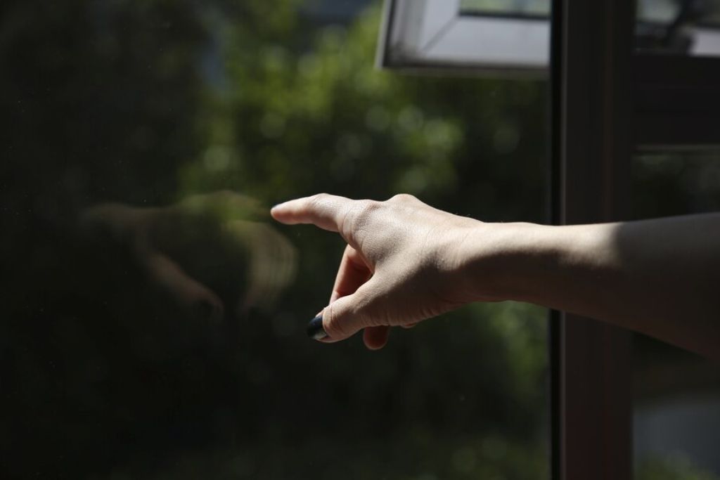 A close-up of a person's hand pointing at a tinted window during the final inspection phase. The dark window film reflects the hand slightly, and greenery is visible through the glass outside. The scene suggests careful attention to detail, checking for imperfections or air pockets.