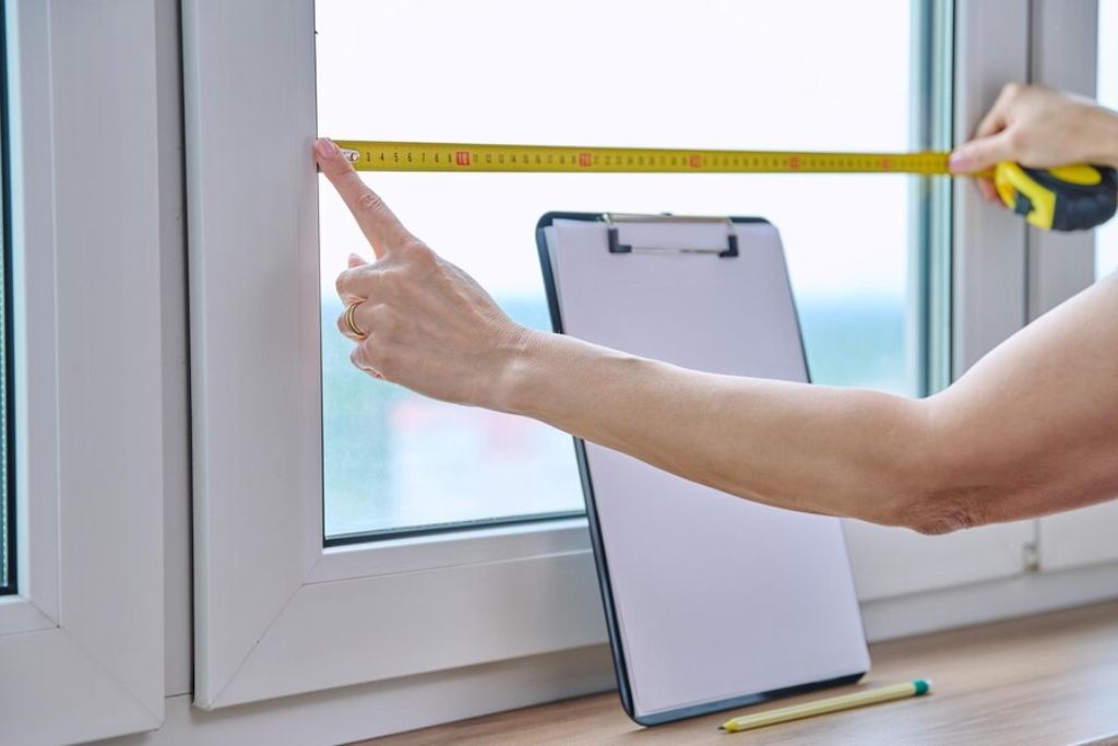 A close-up of a person's hands using a yellow measuring tape to take precise measurements of a window. A clipboard with blank paper and a green pen rest on the windowsill, ready for note-taking. The person's finger points to the measurement mark to ensure accuracy.