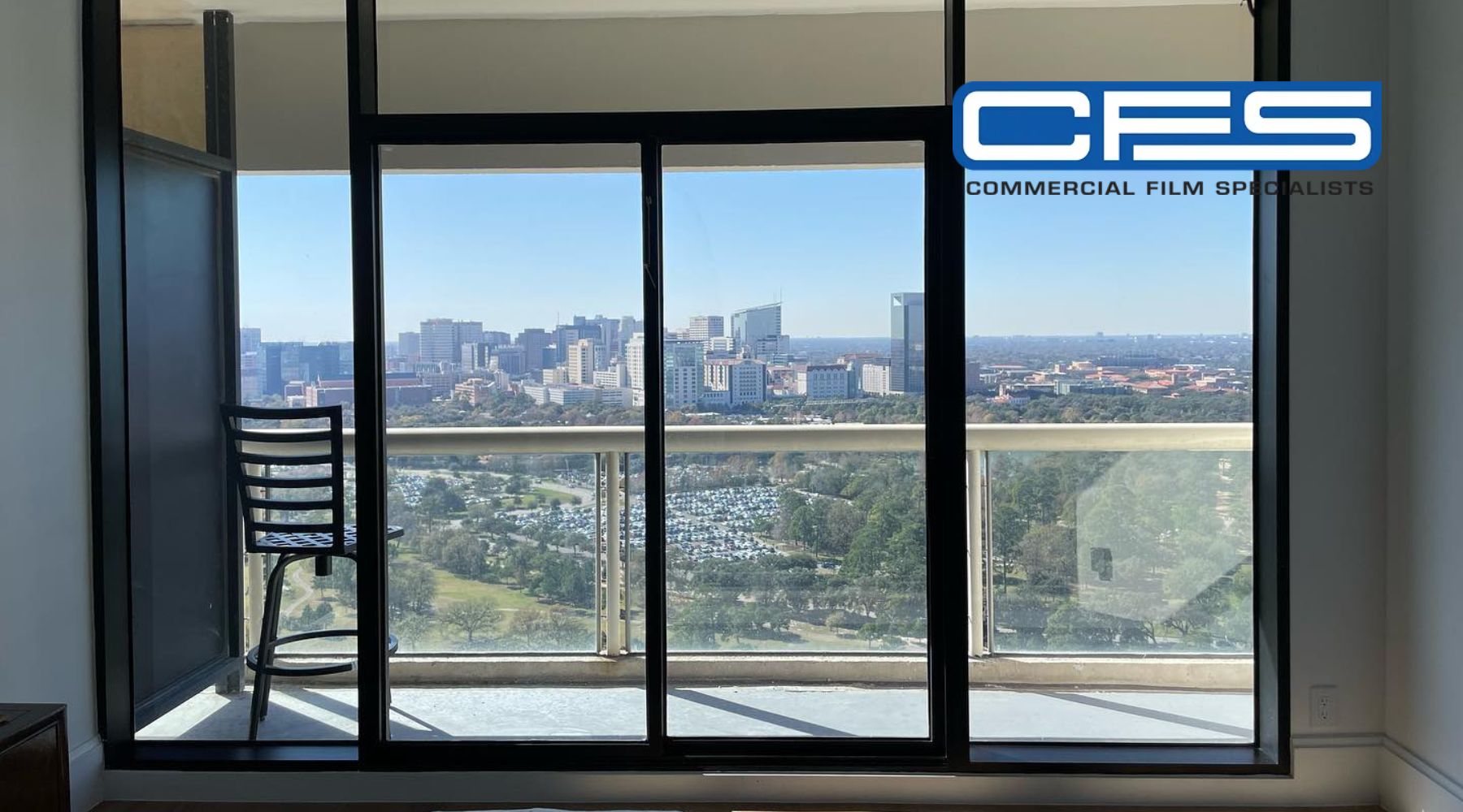 A large glass sliding door with a black frame leads to a balcony overlooking a cityscape with high-rise buildings and green spaces. A black metal chair sits on the balcony, partially visible through the glass. The logo for "CFS - Commercial Film Specialists" is displayed in the upper right corner.