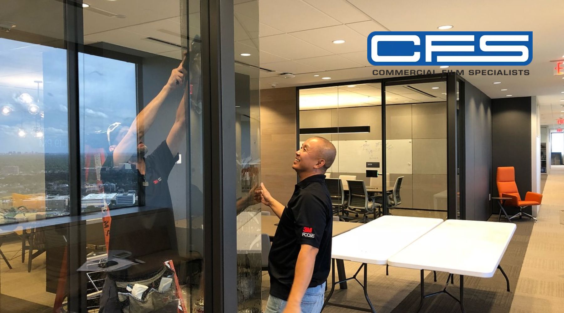 A professional installer from Commercial Film Specialists (CFS) applies window film to an interior glass wall in a modern office setting in Houston, Texas. The installer, wearing a black 3M-branded shirt, carefully smooths the film while using tools to ensure a clean finish. The office features glass partitions, contemporary furniture, and a bright, organized workspace.