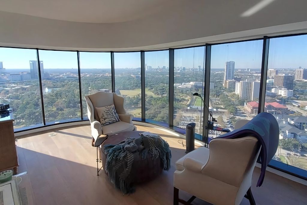 Modern high-rise sitting area with floor-to-ceiling tinted windows curving around the room, offering a panoramic city view. Two armchairs, a lamp, and a cozy throw blanket create a relaxed vibe while tinted glass filters the bright sunlight.