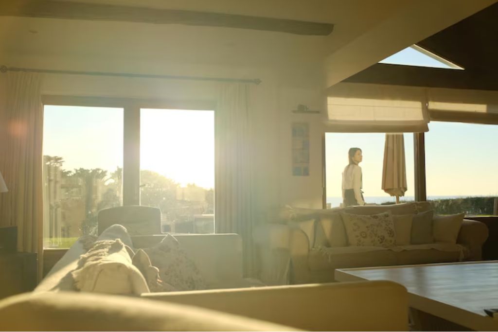 A sunlit living room with large windows and sliding doors letting in intense natural light. Soft, neutral-toned furniture fills the space, while a woman stands outside, bathed in golden sunlight—emphasizing how UV rays can affect interiors over time.