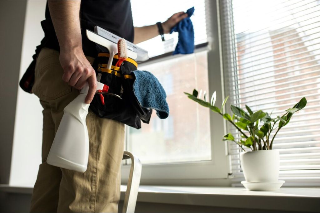 A person stands in front of a window holding a spray bottle and microfiber cloth, with a tool belt stocked with cleaning supplies, as they wipe the tinted window using proper technique. A small potted plant sits on the windowsill.