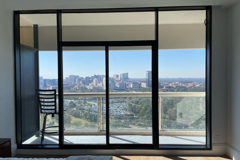 View of a city skyline through large glass sliding doors and windows from inside a high-rise apartment. A black metal chair sits on the balcony, and bright natural light fills the space, highlighting the importance of UV window film.