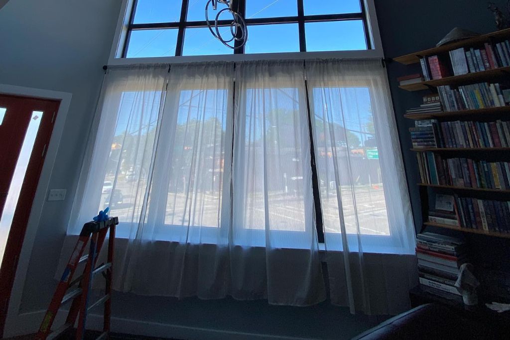 Bright living room with sheer curtains covering large front-facing windows that appear to have window film applied, paired with a tall bookshelf and red ladder nearby, illustrating how layering films and curtains enhances insulation and aesthetic appeal.