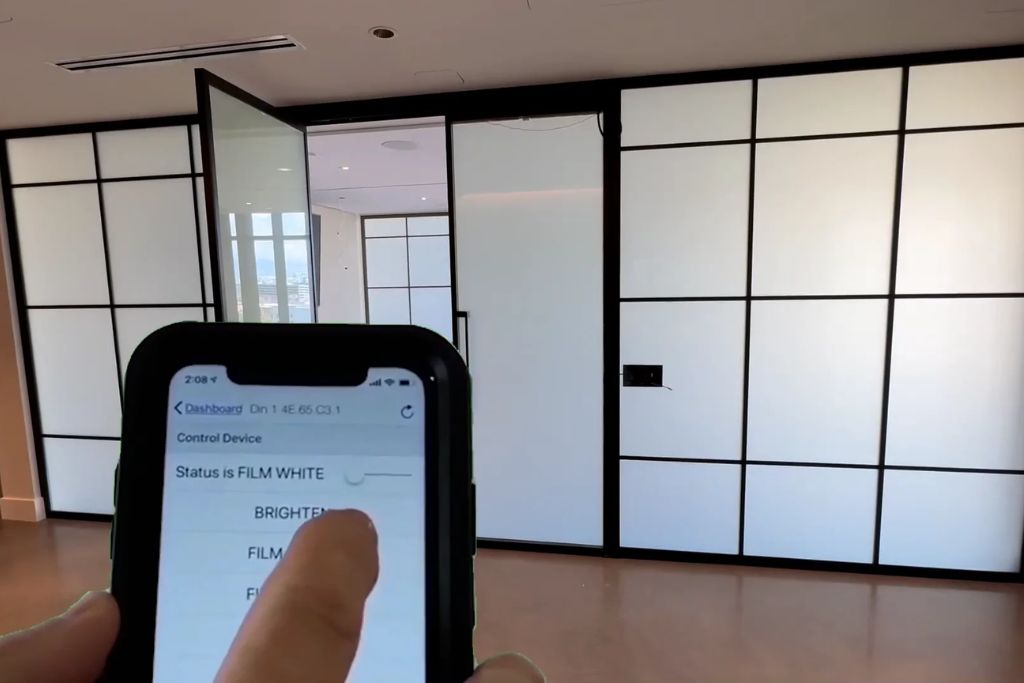 A person uses a smartphone app to control smart window film in a modern room with frosted glass panels that can change opacity. The app screen shows the film status as “FILM WHITE” with options for brightness adjustment.