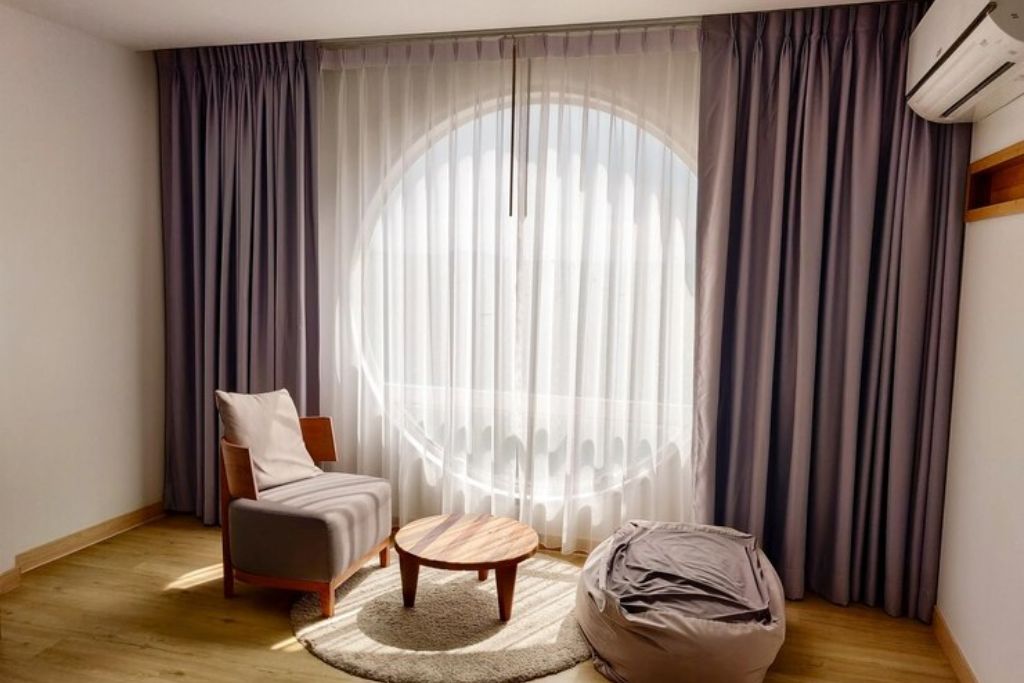 Cozy modern room with a large circular window covered by sheer curtains and thick, pleated thermal curtains in a muted gray tone, designed to insulate against heat and cold while adding a stylish, minimalist touch to the space.