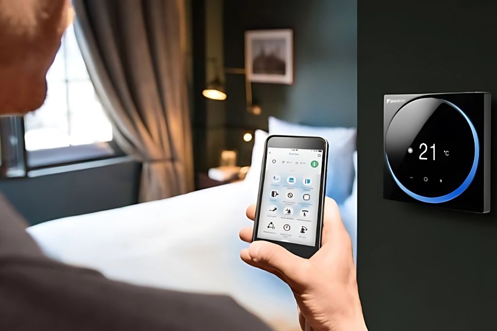 A person uses a smartphone to adjust a smart thermostat in a modern bedroom, with the wall-mounted device displaying a temperature of 21°C. The phone screen shows various smart home control icons, indicating an integrated system.