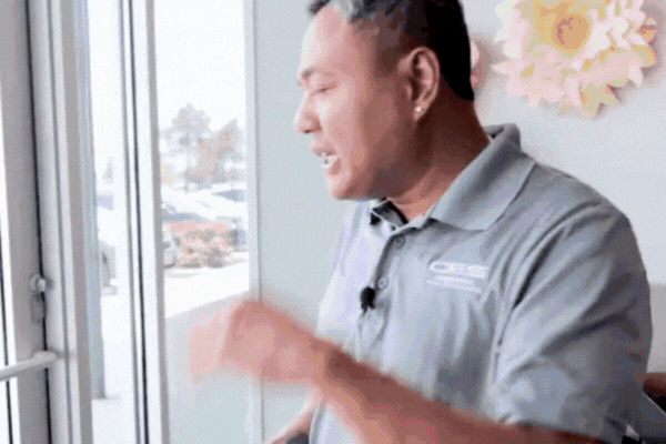 A man in a branded gray polo shirt stands by a large window, gesturing as he explains something, with sunlight streaming in. The window reflects outdoor scenes, highlighting the effectiveness of window film in reducing heat and glare indoors.