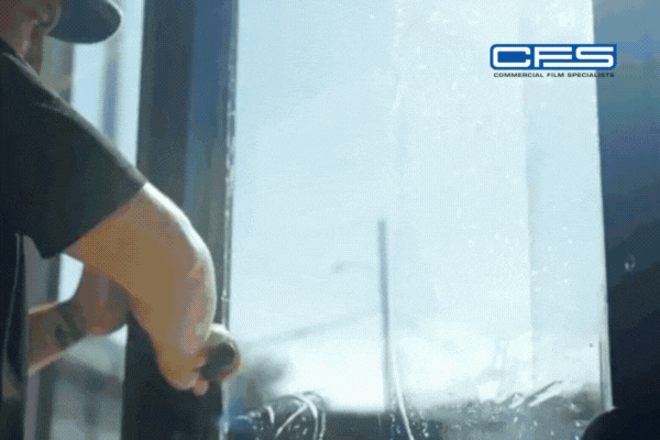 Close-up of a technician applying window film to a large glass pane using a squeegee, with the CFS (Commercial Film Specialists) logo in the top right corner. The film smooths out as the tool passes over it, showing the precision of the installation process.