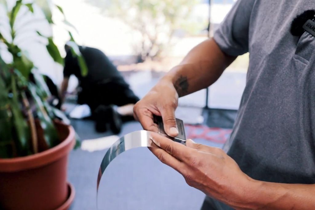 Close-up of a technician’s hands holding a flexible strip of security window film, with another installer working in the background near a window. The scene highlights the material’s clarity, flexibility, and the importance of professional installation.