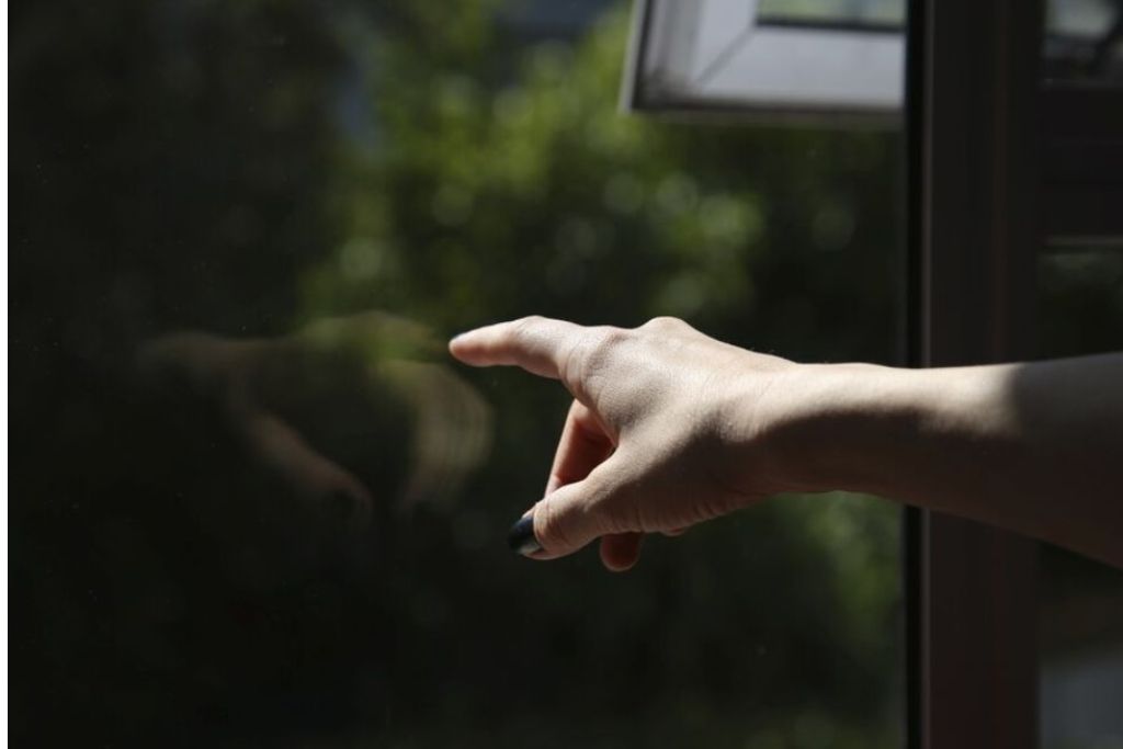 A hand points toward a dark tinted window with greenery faintly visible outside, illustrating how window film can affect visibility and privacy. The reflection on the glass emphasizes its protective and light-controlling qualities.