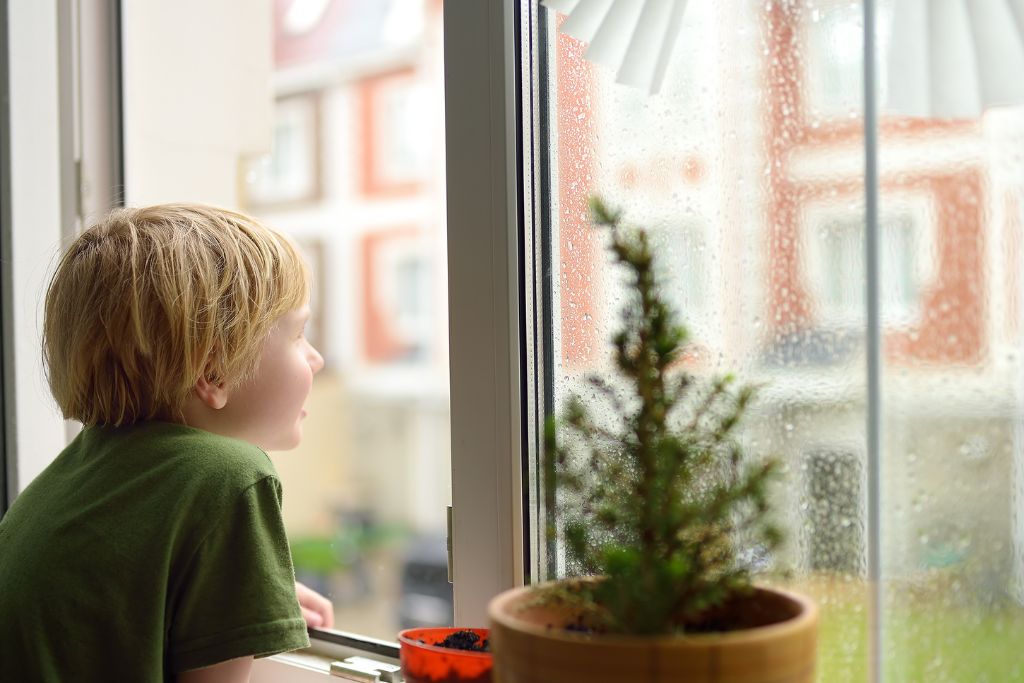 A young child looks out a window covered in raindrops, with a small potted plant on the sill and blurred buildings visible outside. The scene highlights the vulnerability of untreated glass to both weather and security risks.