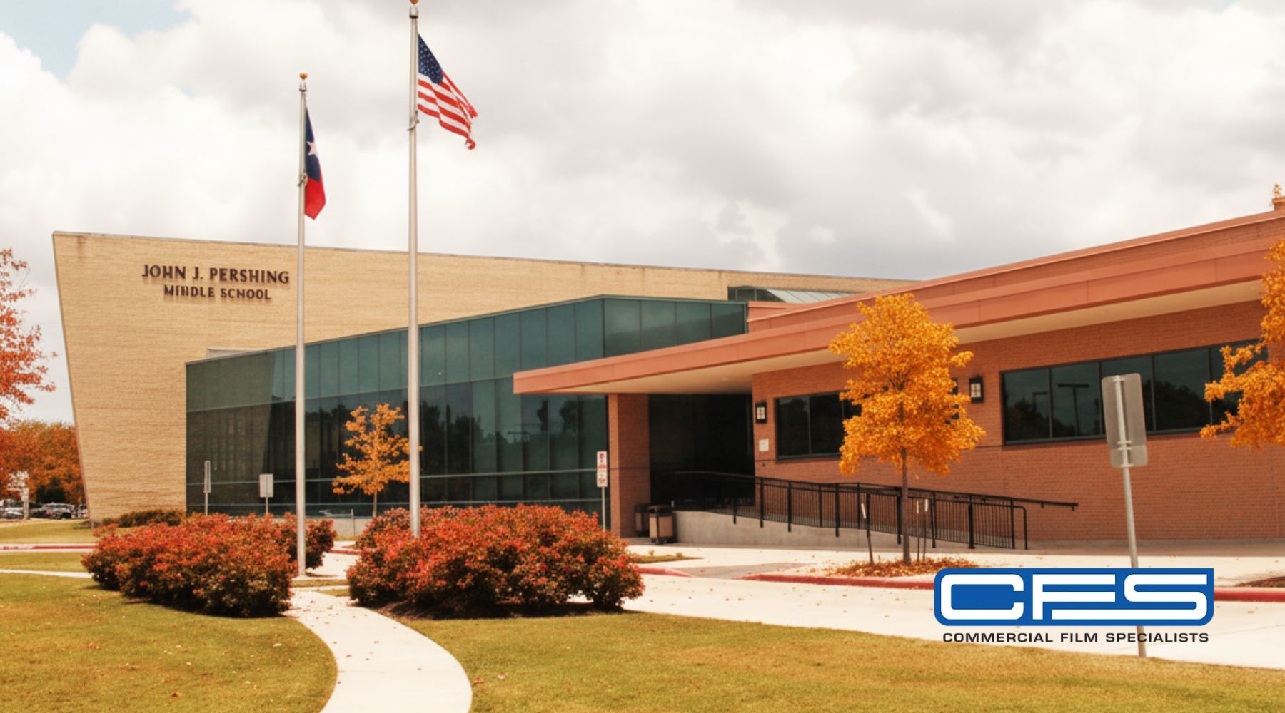 John J. Pershing Middle School in Houston with large glass windows, U.S. and Texas flags flying, and autumn trees with orange leaves in the foreground. The building’s wide glass areas emphasize the need for added security and privacy window tinting.