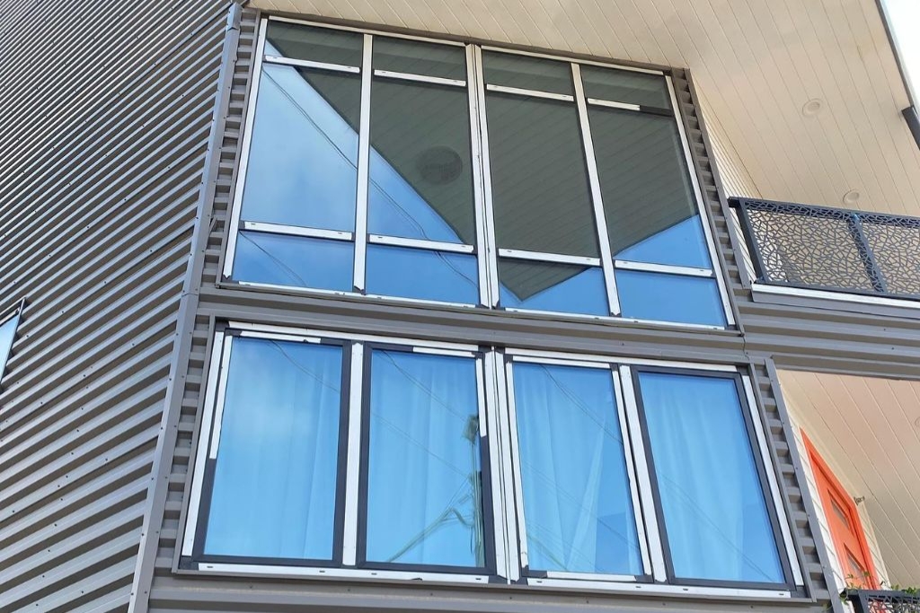 Exterior view of a modern Houston home with large tinted windows framed by corrugated metal siding and a partial view of a balcony. This image demonstrates the benefits of window tinting for enhancing privacy and curb appeal without sacrificing natural light.