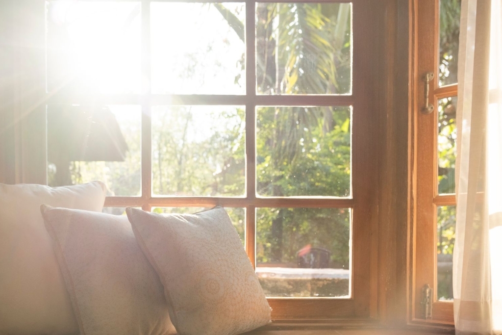 Sunlight pours through a large wooden-framed window with soft cushions on the windowsill, casting a warm glow across the room. This image illustrates how window tinting works by reducing glare and UV exposure while preserving natural light and comfort indoors.
