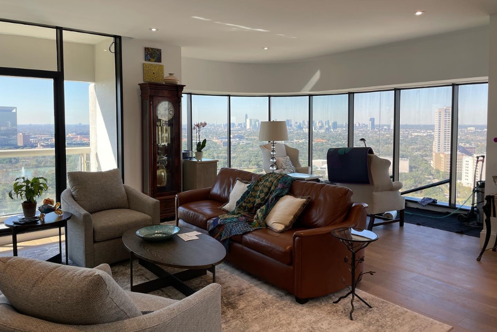 A bright, high-rise Houston living room with floor-to-ceiling windows showcases a panoramic city view, natural light, and elegant furnishings including a leather sofa and tall grandfather clock. This image illustrates why Houston homes benefit most from window tinting, as it helps manage glare, UV exposure, and temperature in sun-filled spaces.