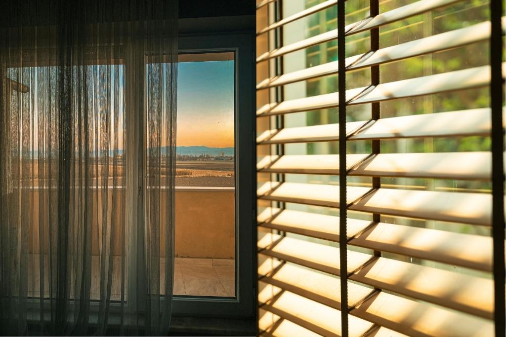 Interior view of a window with sheer curtains and horizontal blinds, showing warm sunlight and an outdoor landscape at sunset.